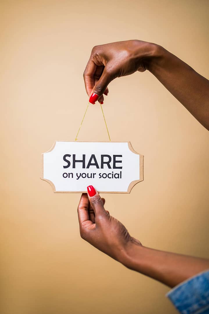 Close-up of a person's hand holding a 'Share on your social' sign against a beige background.